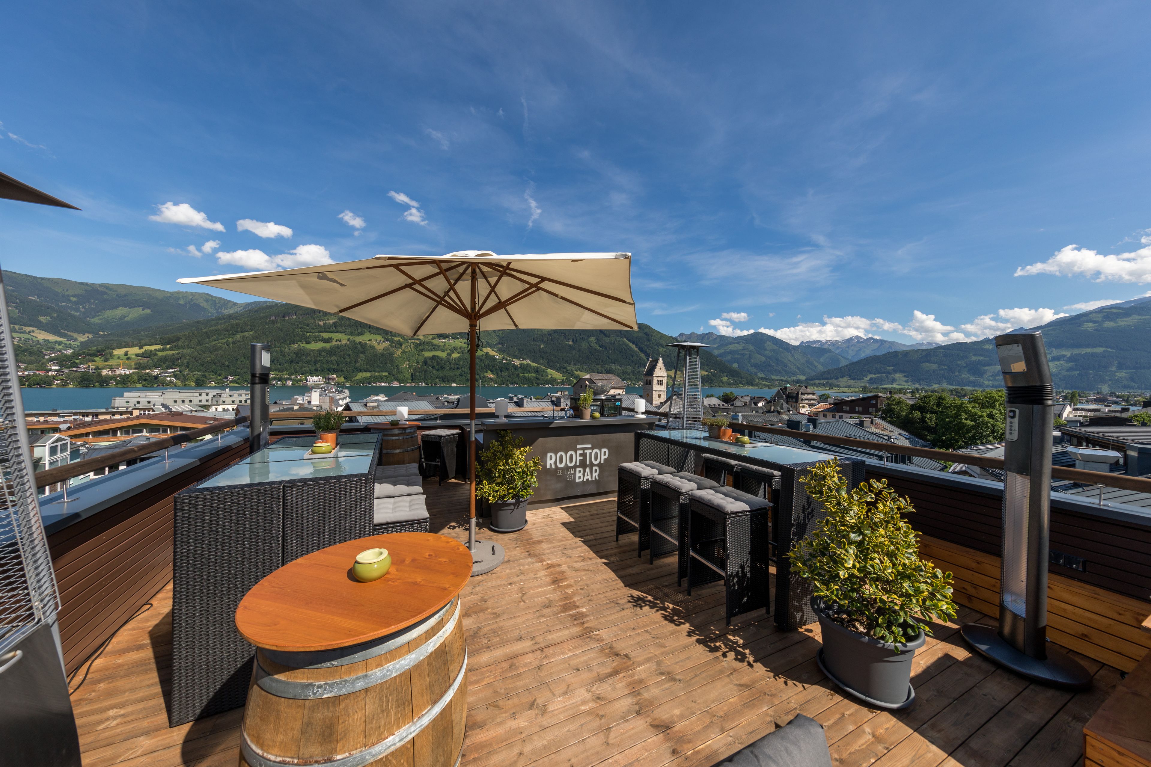 A rooftop bar with outdoor seating, a large umbrella, and a view of the mountains and lake.