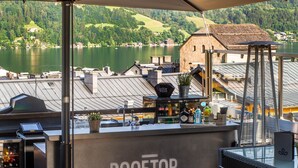 A rooftop bar with a view of a lake and buildings in the background. - HEITZMANN - Hotel & Rooftop (Zell am See)