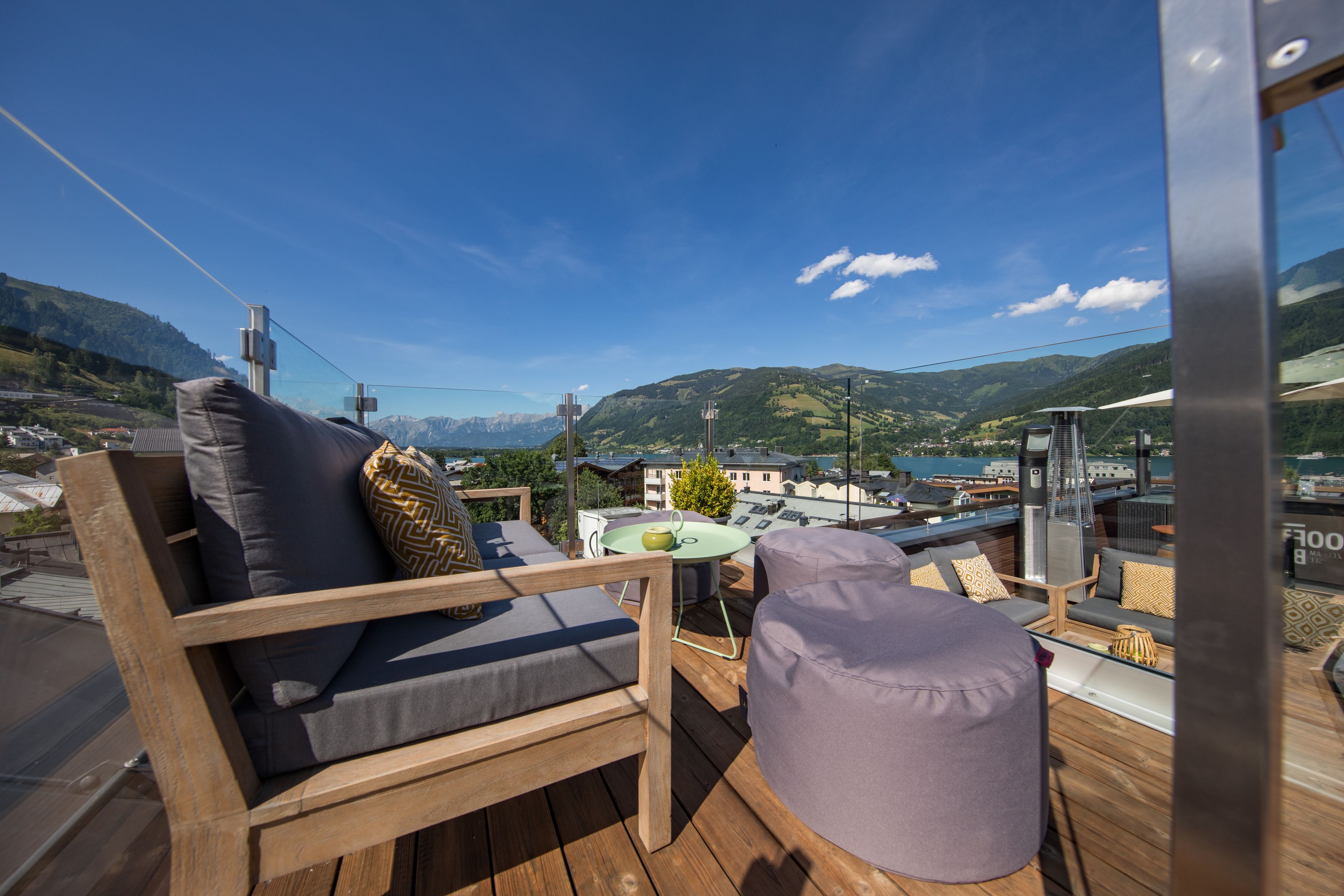 Une terrasse en bois avec un canapé et un pouf, une petite table avec un plateau vert, et une vue sur un paysage montagneux.