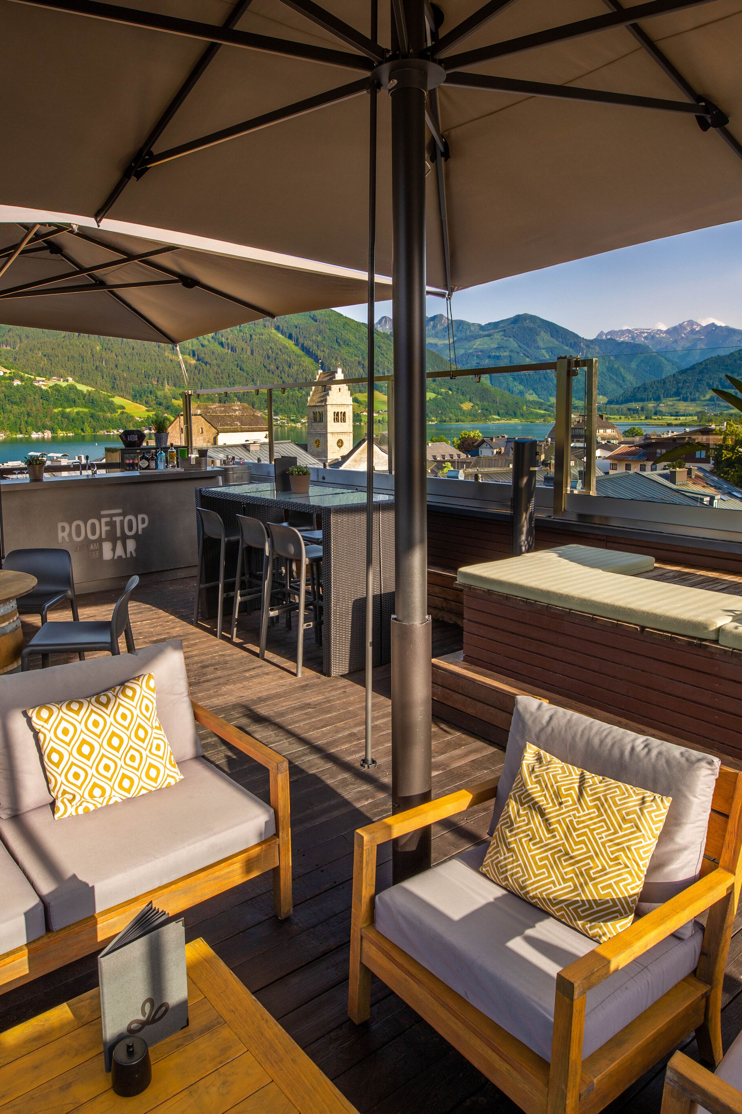Eine Dachterrasse mit Außenbestuhlung, einer Bar und Blick auf Berge und See.
