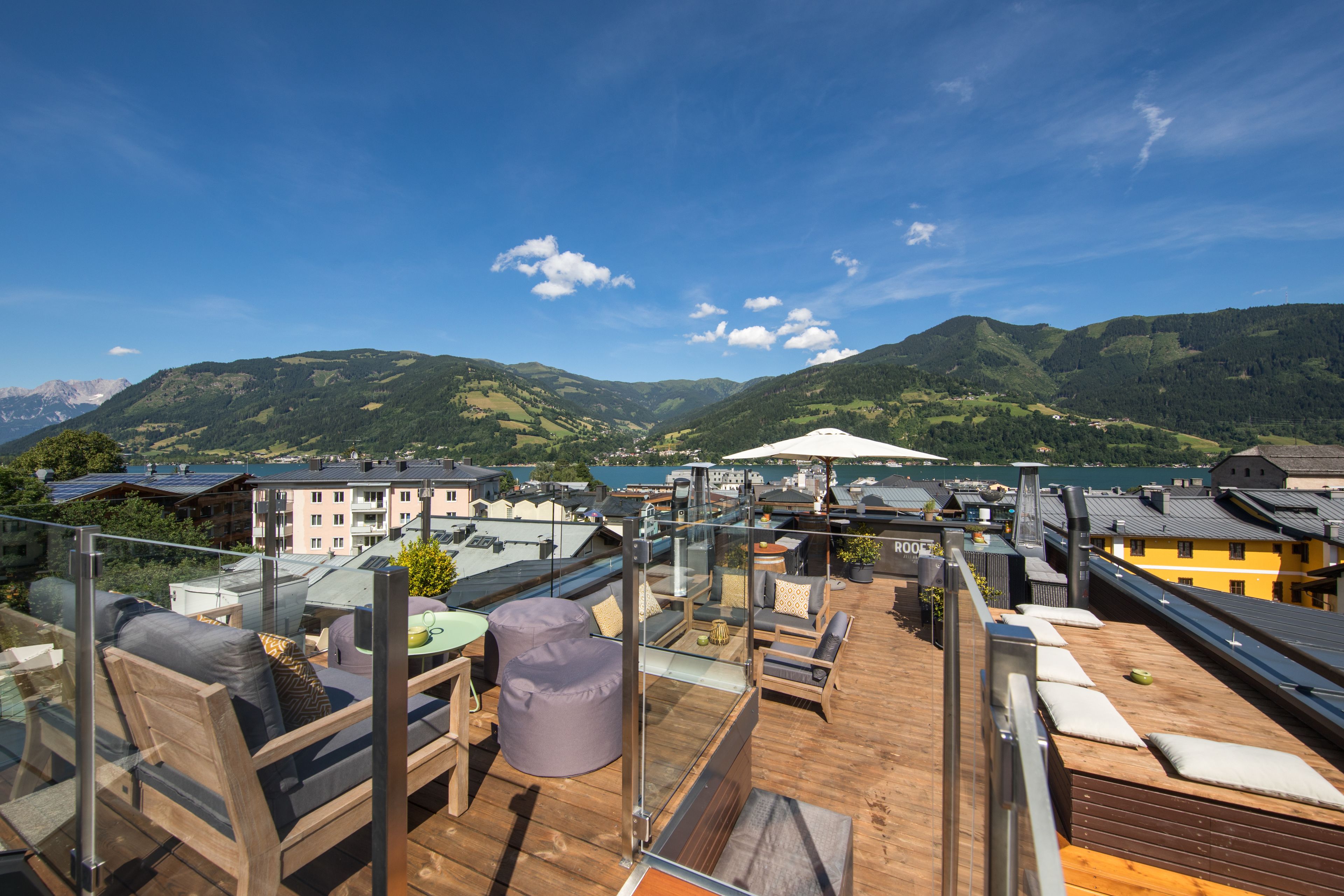 A rooftop terrace with outdoor seating, a glass balustrade, and a view of mountains and a lake.