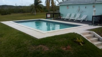 Una piscina rectangular con borde de azulejos, rodeada por un césped. Hay sillas de descanso blancas alineadas frente a un edificio azul claro con techo.