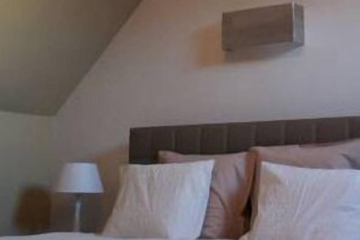 A bedroom with a sloped ceiling, a bed with white bedding, a nightstand with a lamp, and a wall-mounted air conditioning unit.