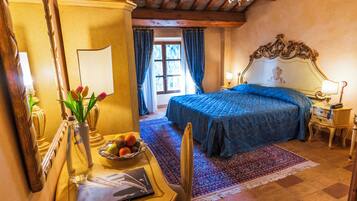 A bedroom with a large bed, a desk with a vase and fruit bowl, a mirror, and a chandelier.