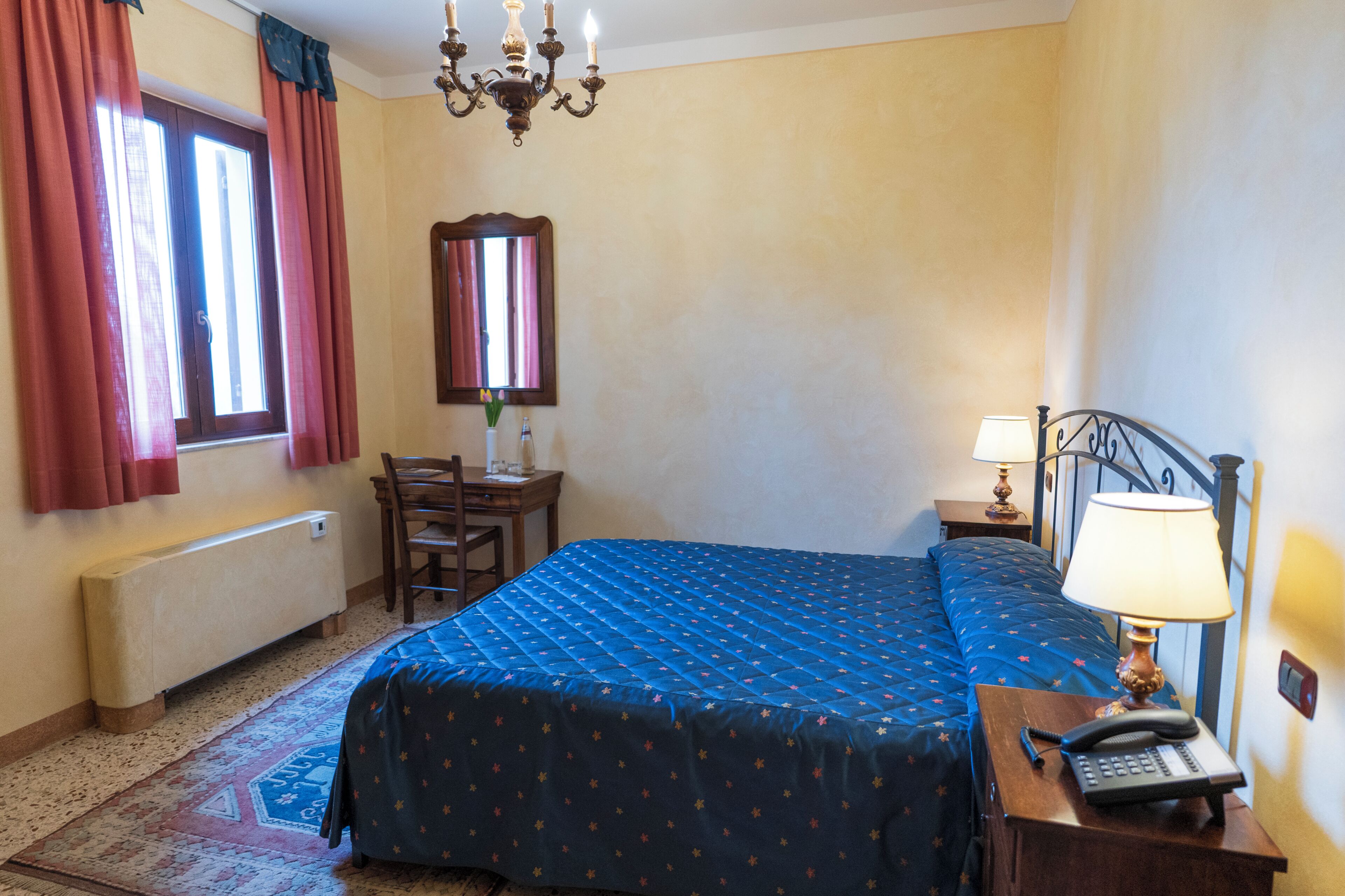 Un dormitorio con cama, escritorio, silla, mesita de noche, lámpara y espejo.