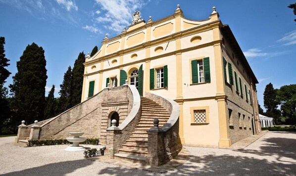 A large, yellow building with green shutters, a stone staircase, and a fountain in front.