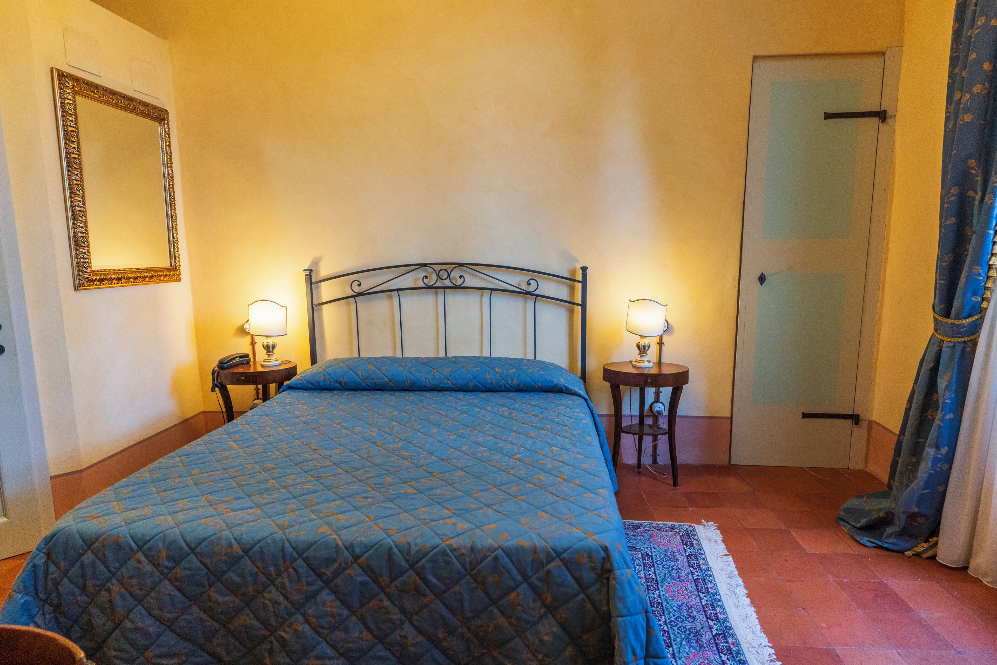 A bedroom with a bed, two bedside tables with lamps, a mirror, a door, and a patterned rug.