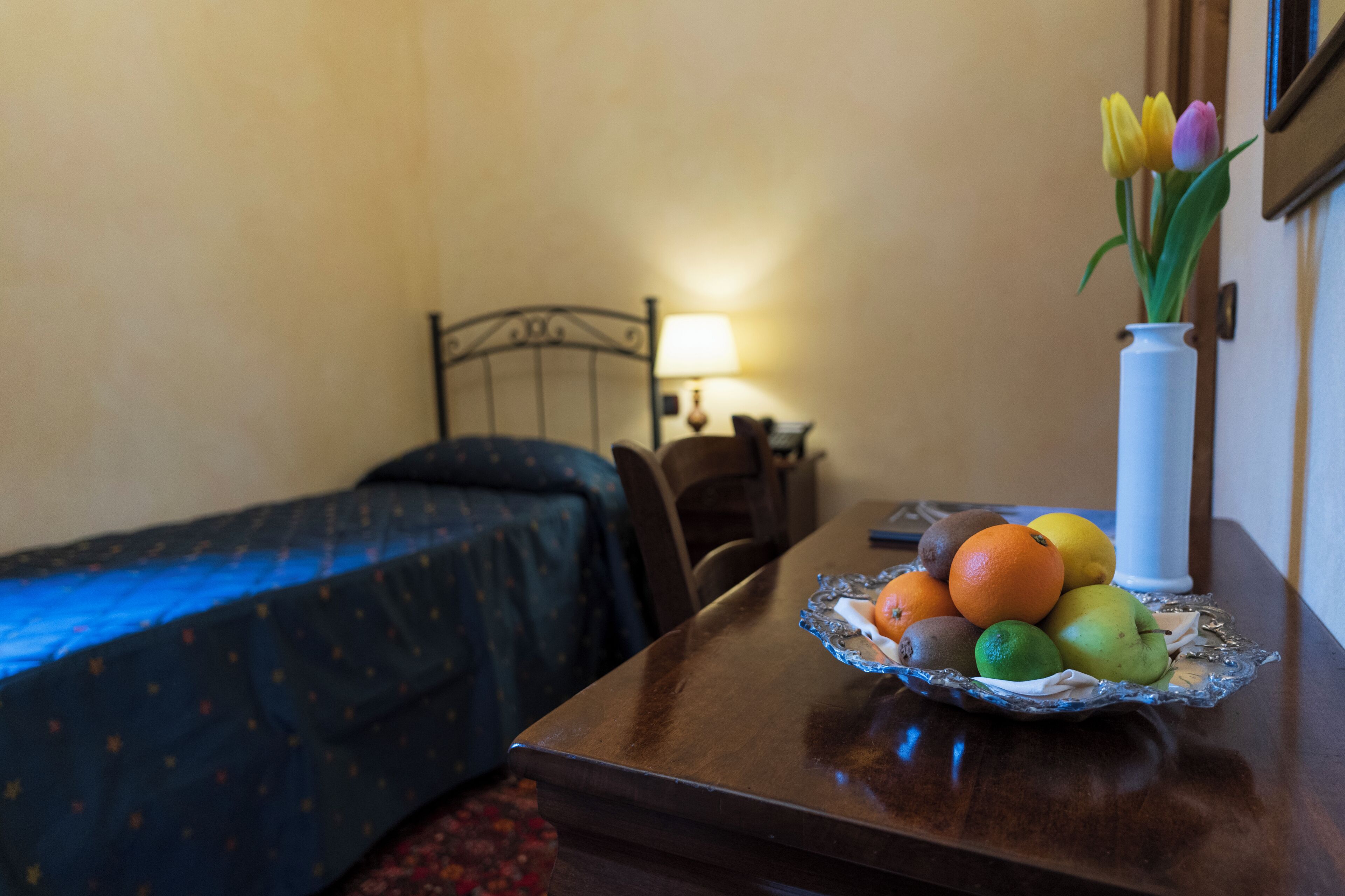 Quarto de hotel com cama, mesa de cabeceira com abajur, e uma mesa com frutas e um vaso de flores.