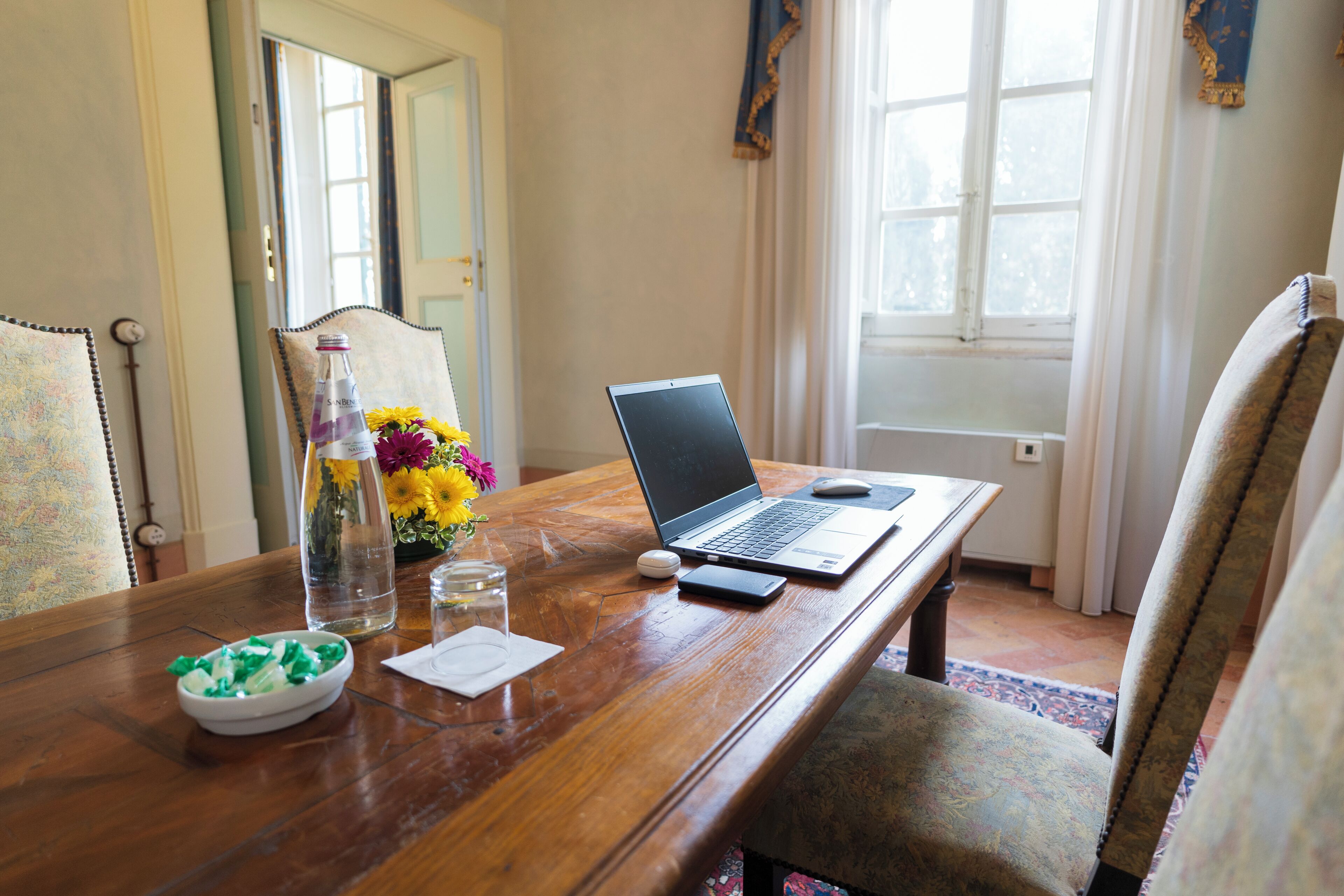Una mesa de madera con una computadora portátil, un teléfono inteligente, un vaso de agua, un florero con flores y un cuenco de aperitivos.