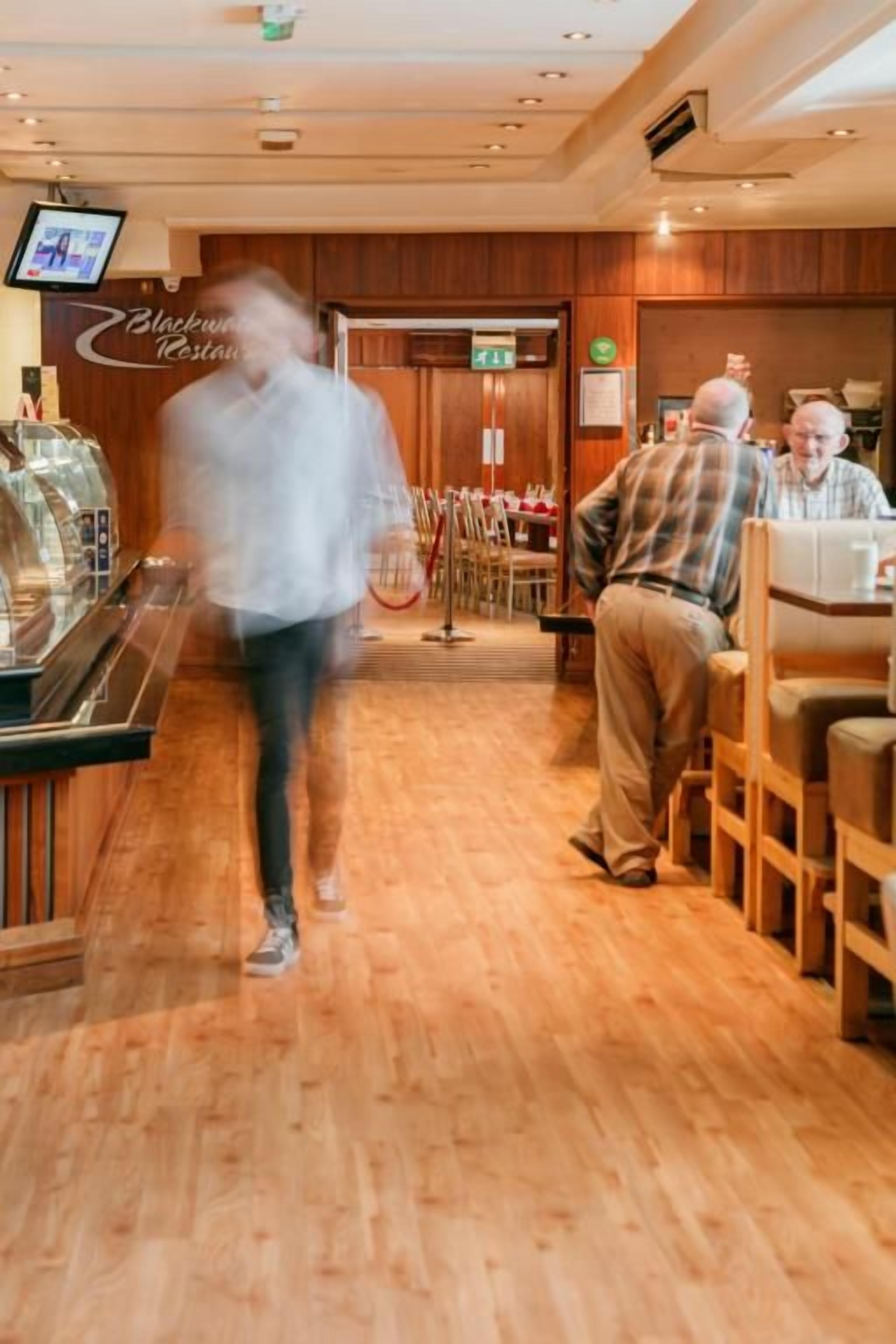 Corredor de hotel com piso de madeira, máquina de café e área de restaurante com mesas e cadeiras.