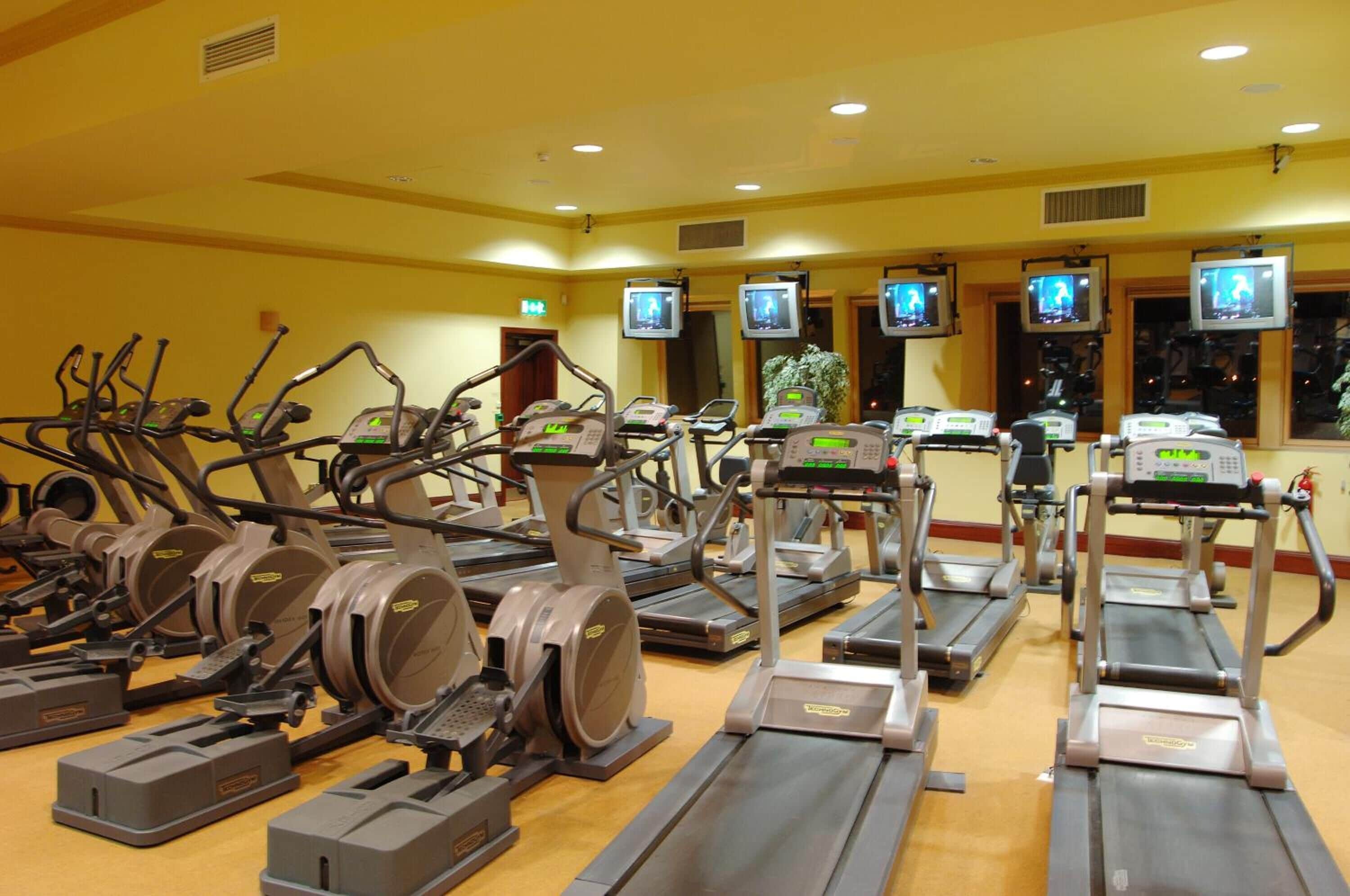 A gym with multiple treadmills and elliptical machines, equipped with digital screens and weight benches.