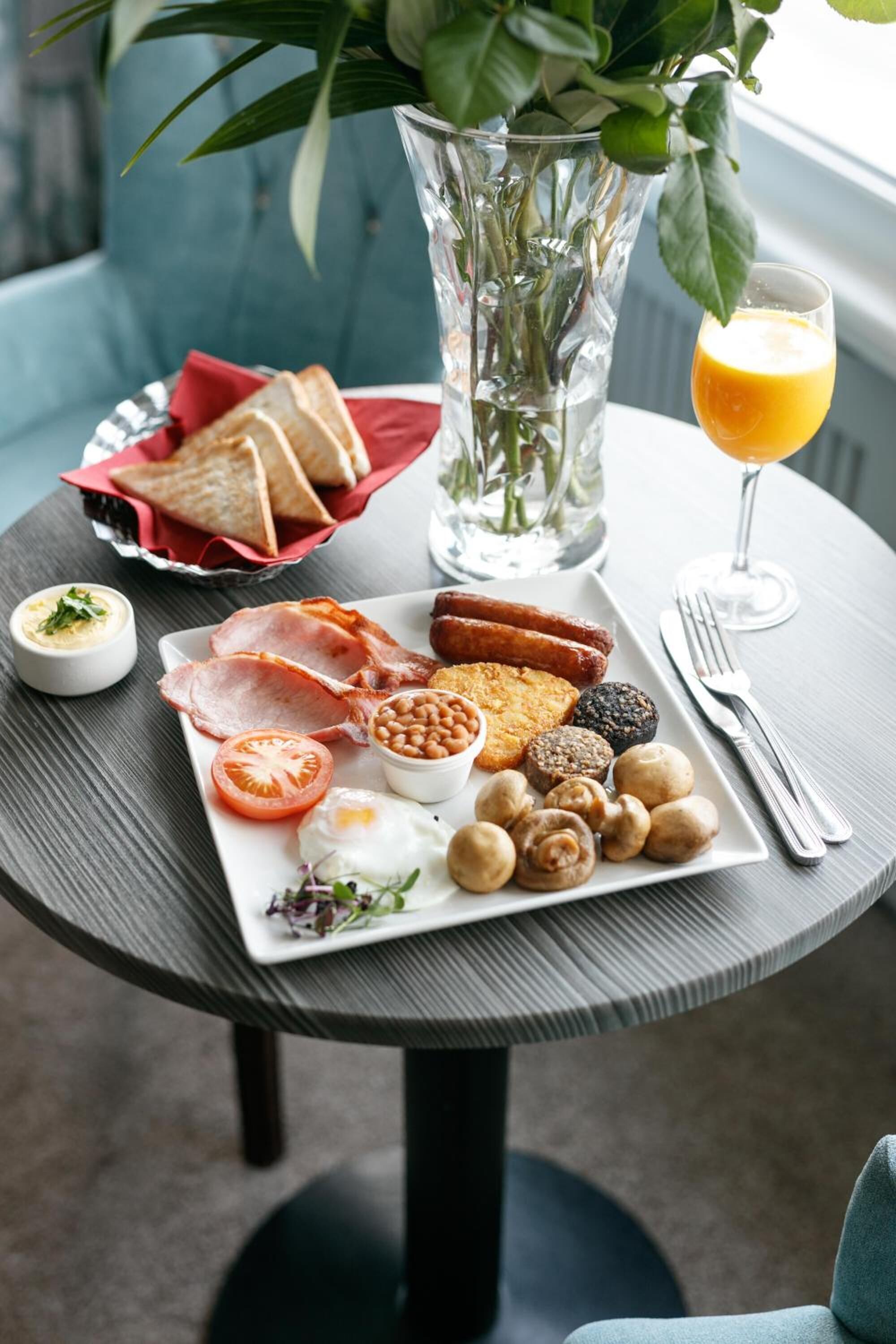A breakfast table with toast, eggs, sausages, beans, mushrooms, and a glass of orange juice.