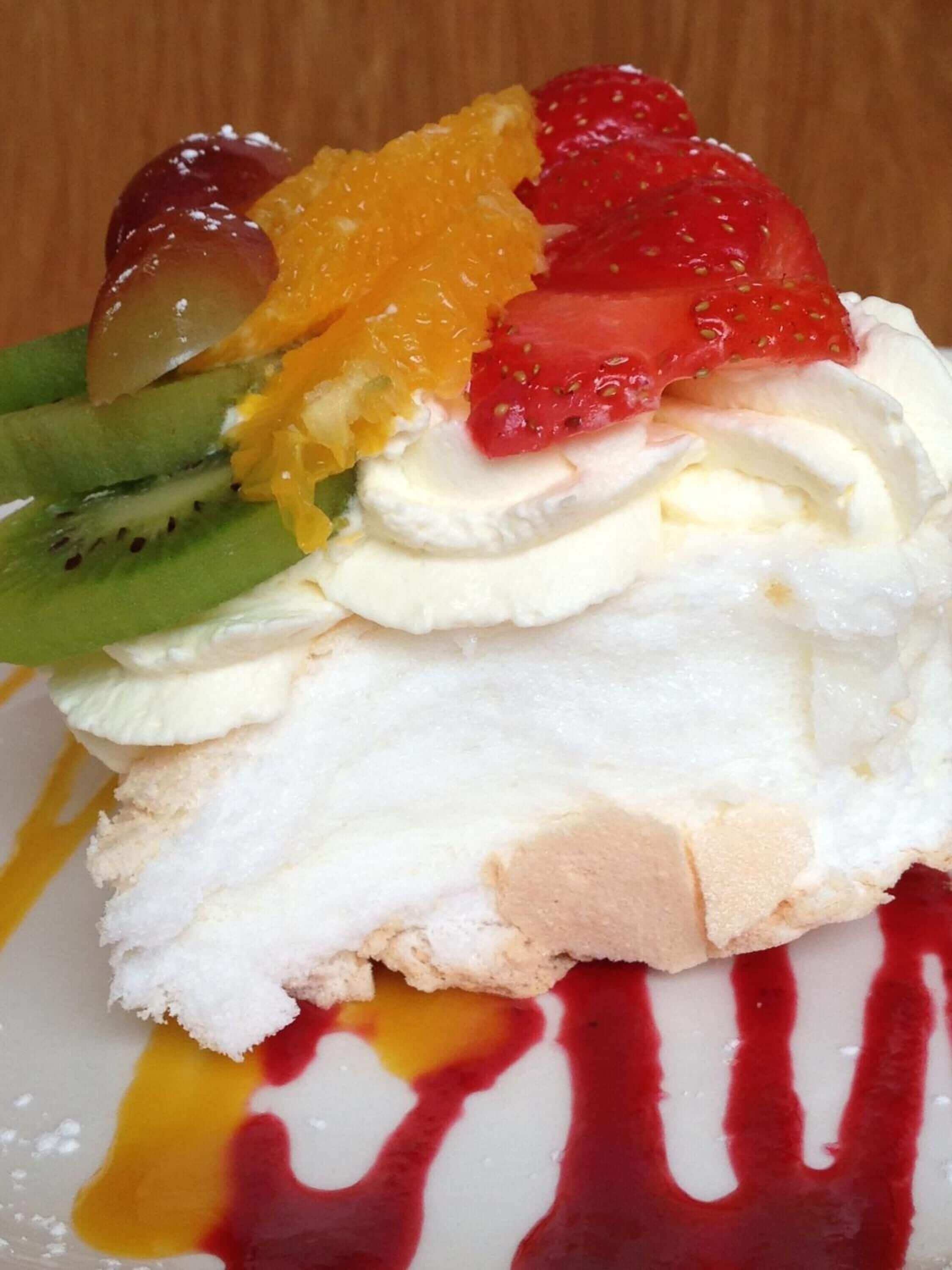 A dessert with whipped cream, strawberries, kiwi, and orange segments on a white plate with red sauce drizzled around.