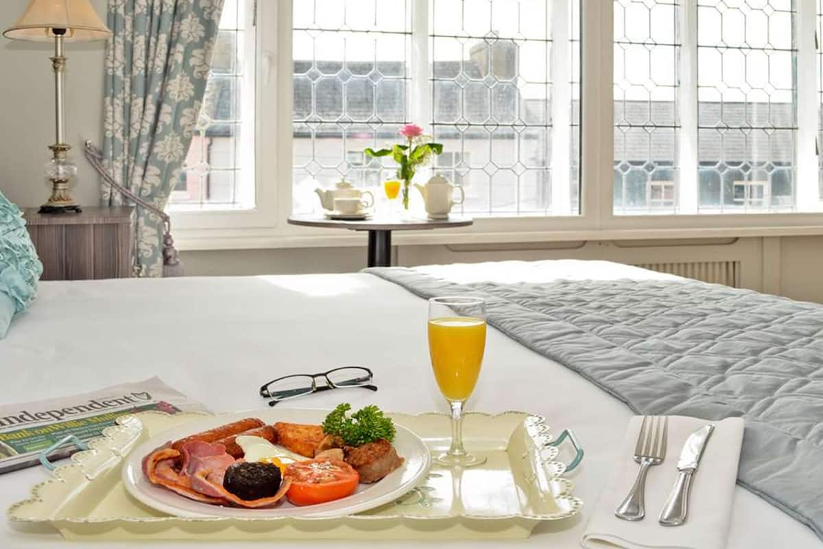 Un lit bien fait, recouvert d’une couette blanche, une table de chevet avec une lampe, un plateau avec des éléments de petit-déjeuner et une fenêtre donnant sur des immeubles.