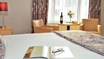 A hotel room with a bed, a bedside table with a lamp, a chair, a small table with a bottle and flowers, and a window with curtains.