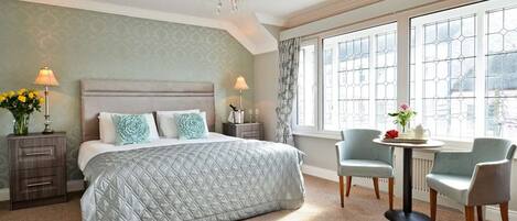 A bedroom with a large bed, two bedside tables with lamps, a chandelier, a window with a view, and two chairs around a small table.