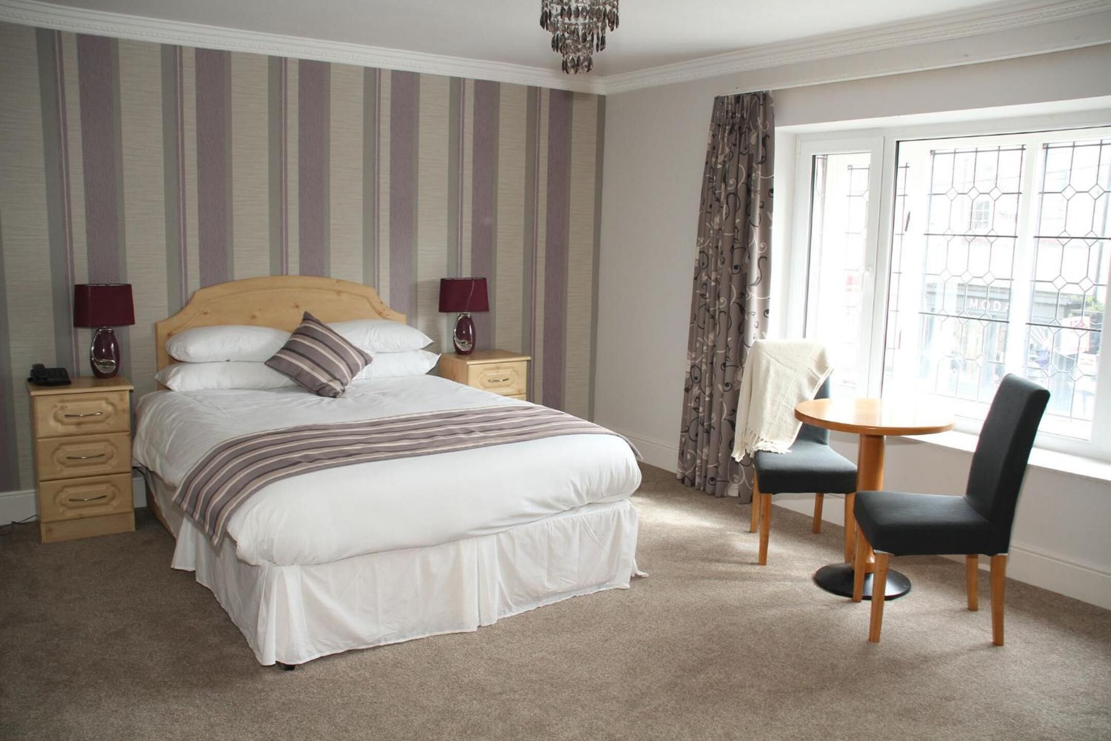 A bedroom with a large bed, two bedside tables with lamps, a small round table with a chair, and a window with curtains.