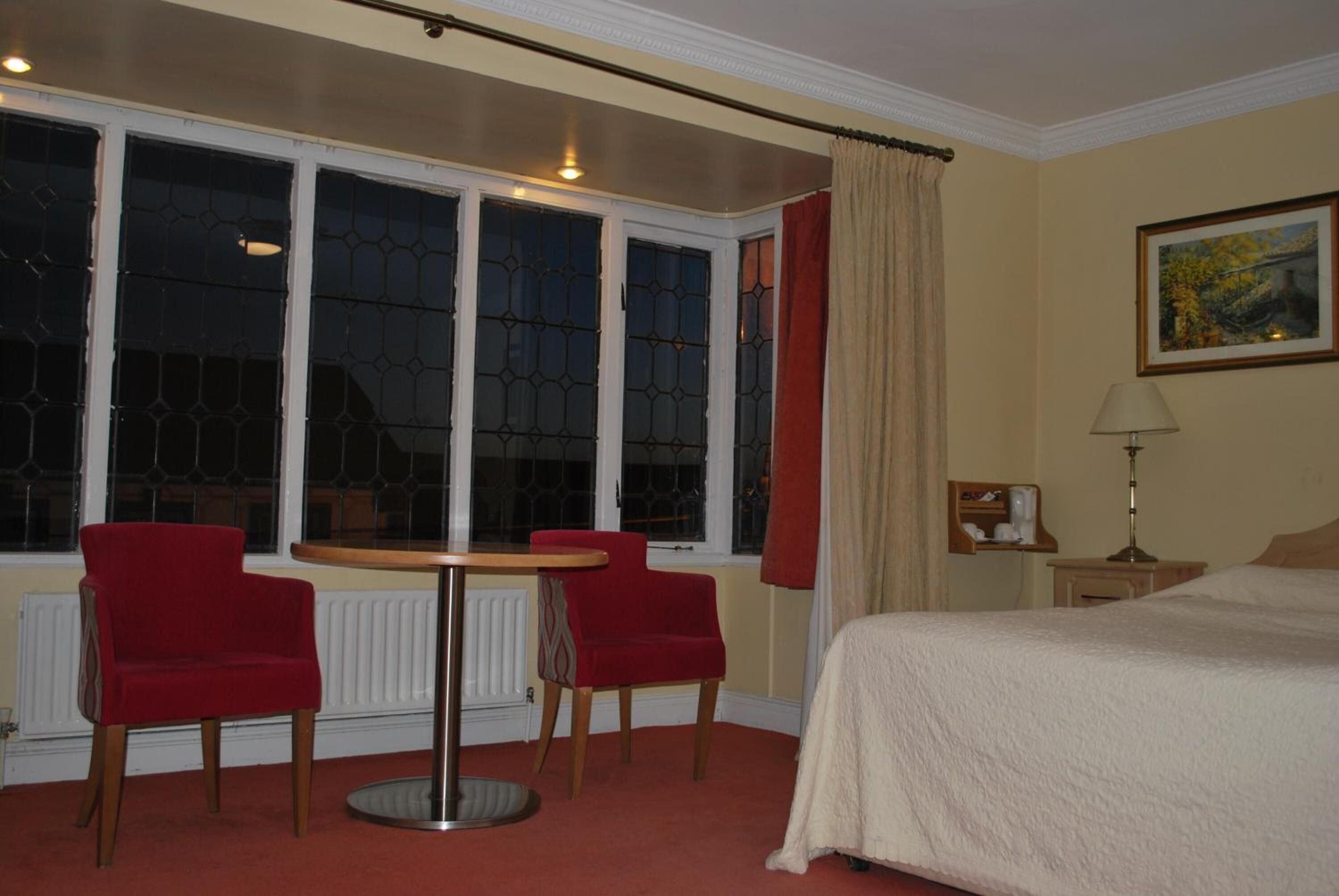 Una habitación de hotel con una cama, dos sillas rojas, una mesa redonda, una ventana con cortinas, un cuadro en la pared y una mesita de noche con una lámpara.