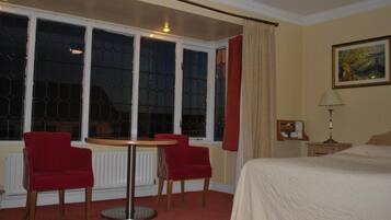 Une chambre d’hôtel avec un lit, deux fauteuils rouges, une table ronde, une fenêtre avec des rideaux, un tableau au mur et une table de chevet avec une lampe.