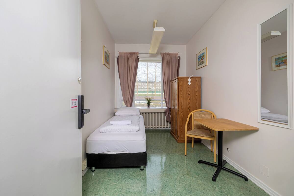 Una habitación pequeña y limpia con cama, escritorio y silla. Tiene una ventana con cortinas, un radiador y una puerta con un cartel.