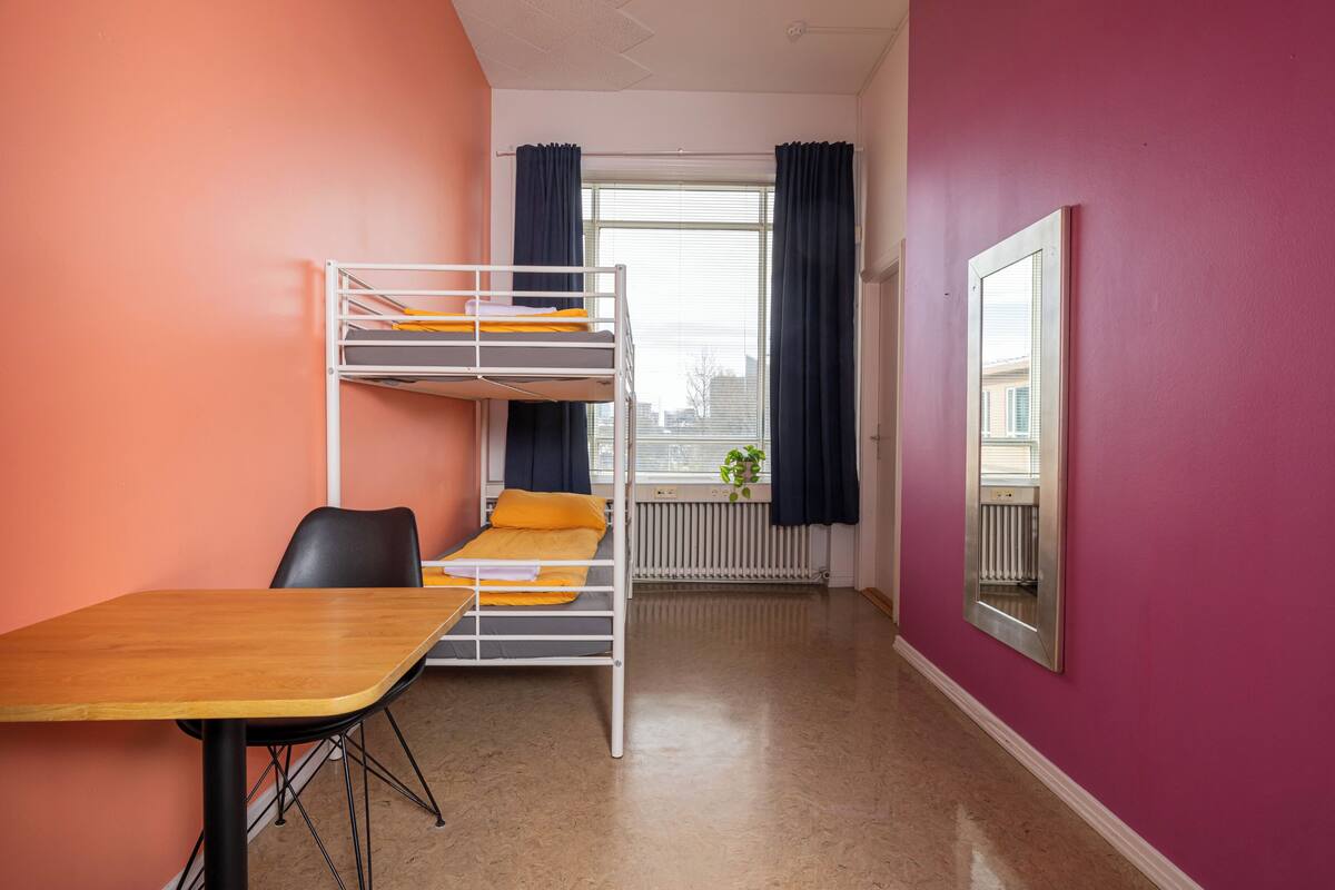 Una cama litera con ropa de cama naranja, una mesa de madera y una silla negra en una habitación con paredes rosas y una ventana con cortinas.