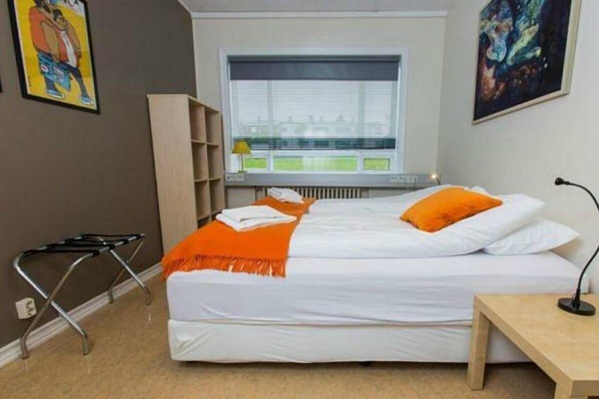 Habitación de hotel con cama, mesita de noche, una silla, ventana con persianas, radiador y cuadros en las paredes.