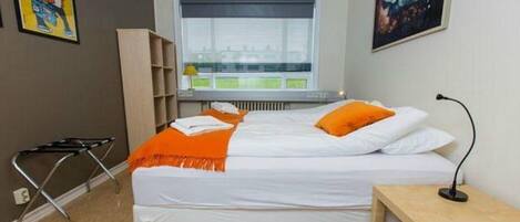Habitación de hotel con cama, mesita de noche, una silla, ventana con persianas, radiador y cuadros en las paredes.