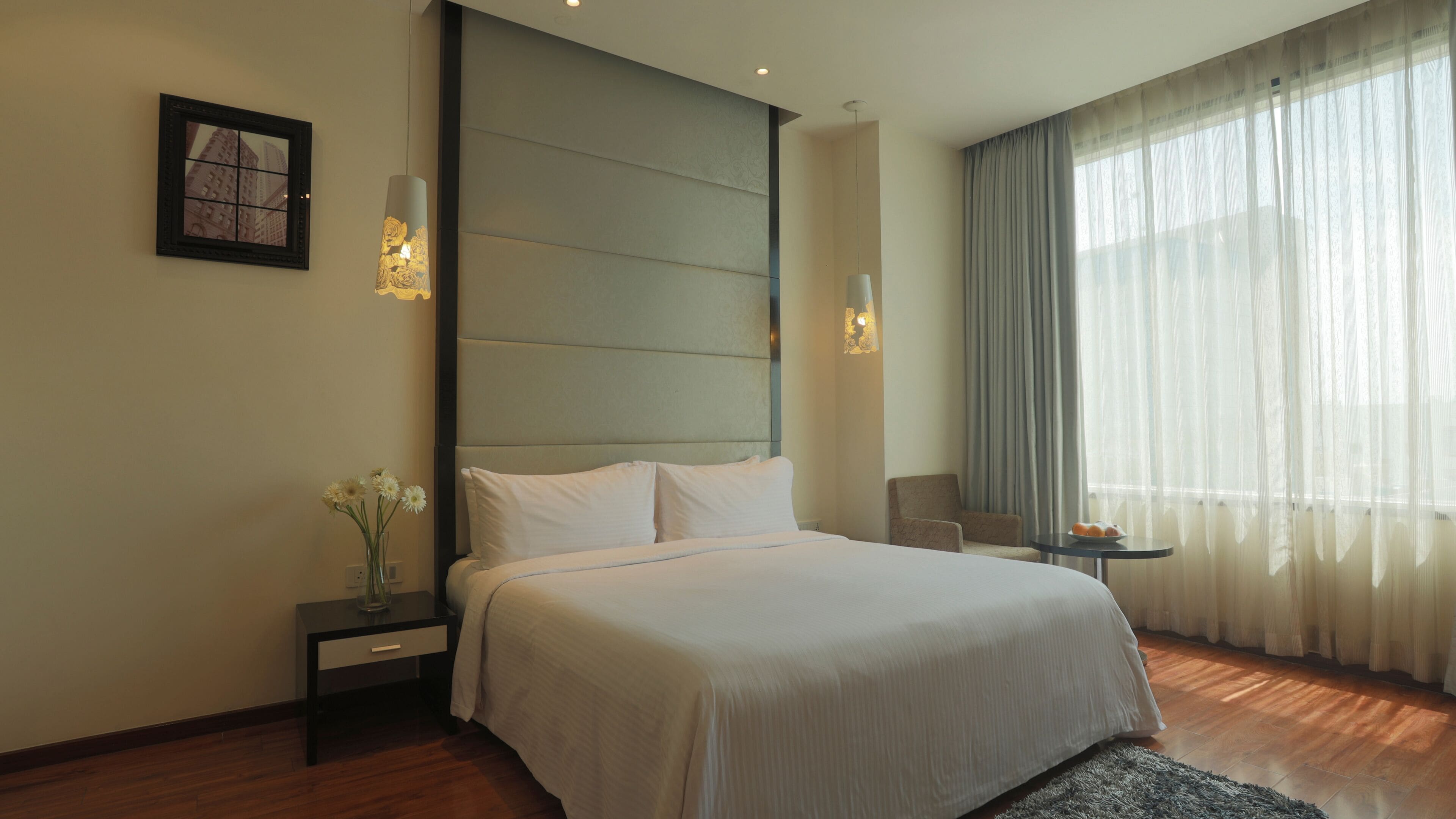 A hotel room with a large bed, a nightstand with a vase of flowers, a chair, and a table with a plate of fruit.