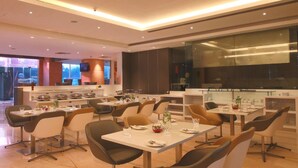 A modern dining area with multiple tables and chairs, a kitchen in the background, and a large window providing natural light. - E Hotel (Chennai)