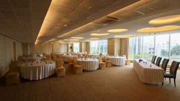 A spacious conference room with multiple round tables, large windows offering a city view, and a high ceiling with ventilation.