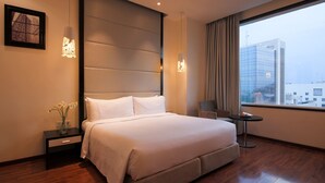 A modern hotel room with a large bed, a nightstand, a vase of flowers, a chair, a small table, and a view of the city through a large window. - E Hotel (Chennai)