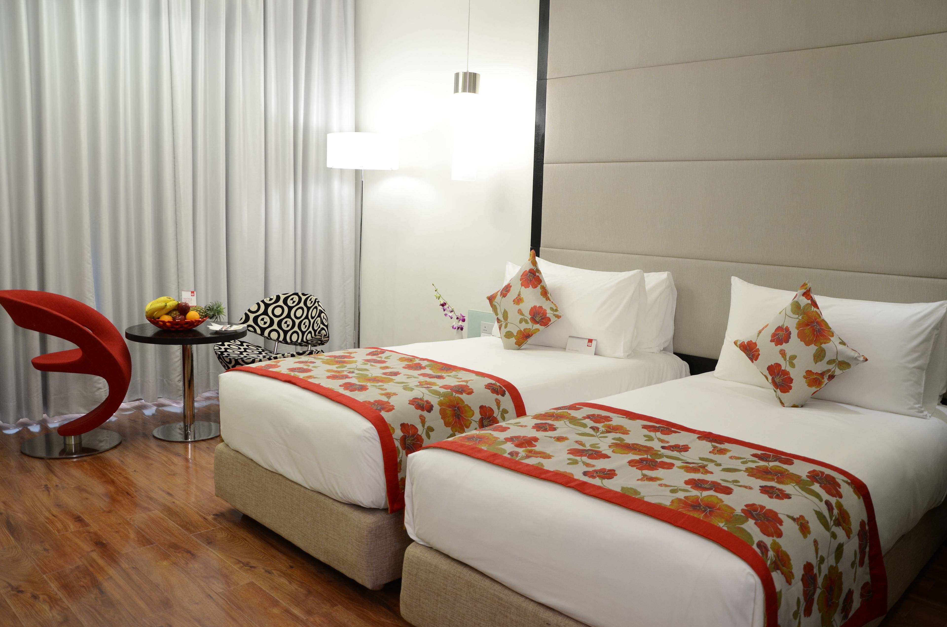 Ein Hotelzimmer mit zwei Betten, einem roten Sessel, einem kleinen Tisch mit einer Obstschale und einer Wanddekoration mit Muster.