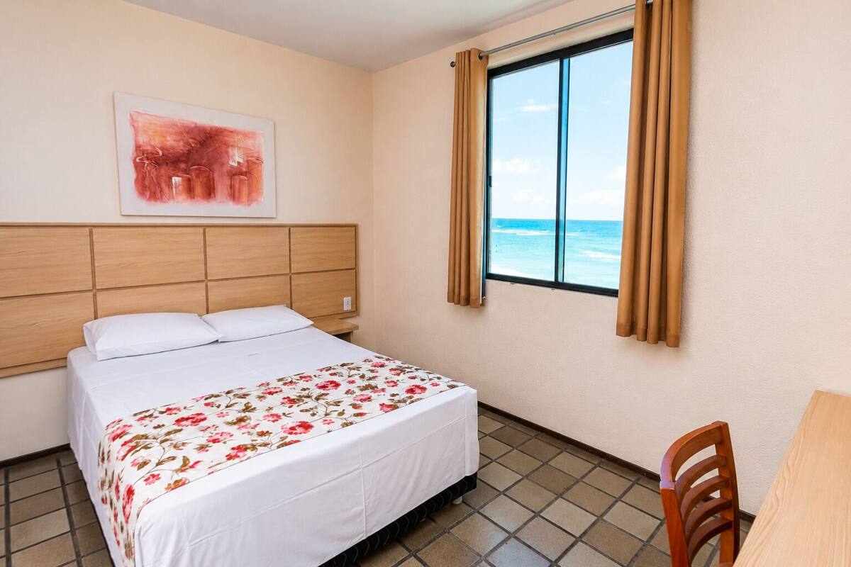 A hotel room with a bed, a window with curtains, a wooden headboard, and a painting on the wall.