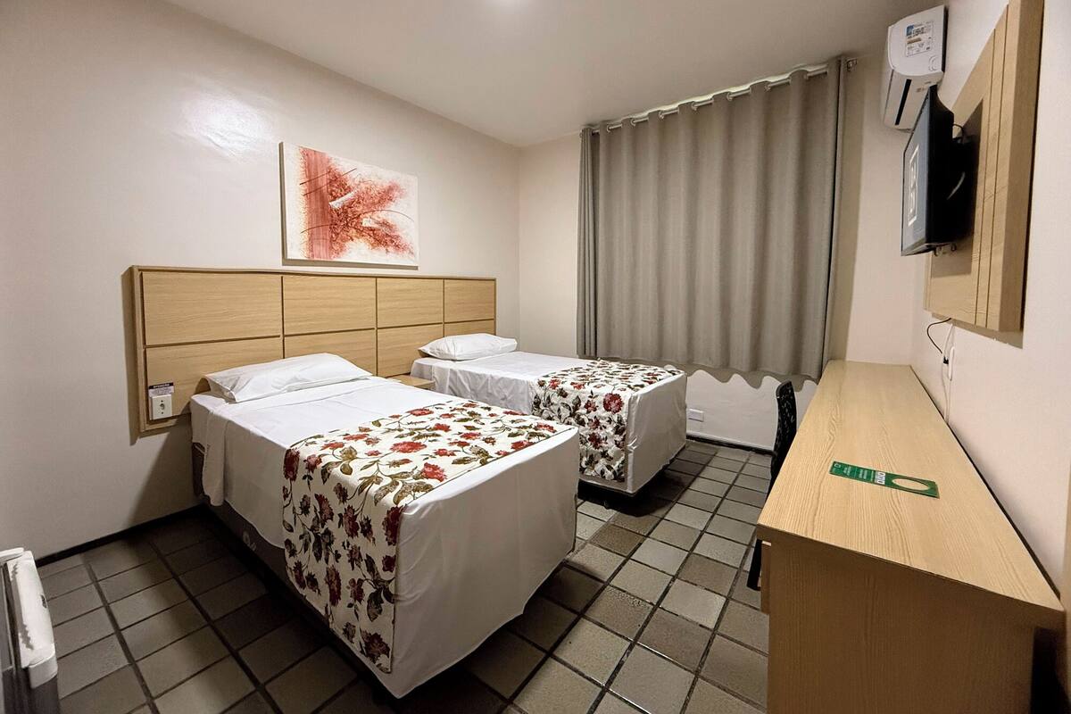 A hotel room with two beds, a wooden headboard, a flat-screen TV, a window with curtains, and a tiled floor.
