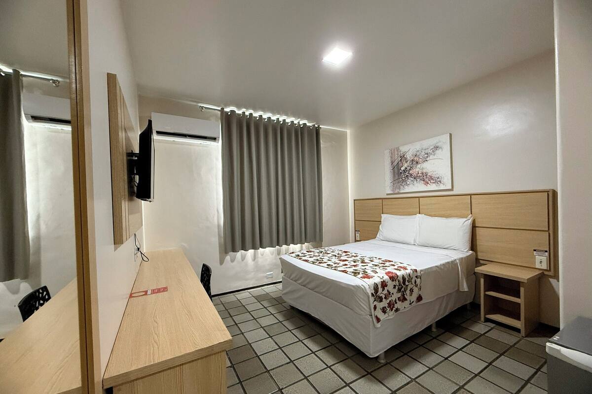 A hotel room with a bed, a desk, a chair, a nightstand, a TV, and a window with curtains.