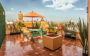 Une terrasse sur le toit, avec un sol carrelé, des chaises en métal vert, une table et un parasol orange.