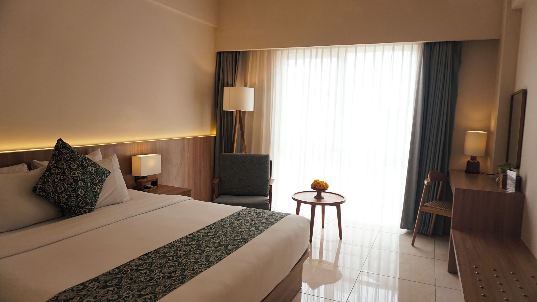 A hotel room with a large bed, a desk, a chair, a small table with a vase, and a window with curtains.