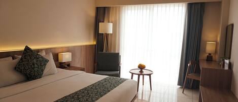 A hotel room with a large bed, a desk, a chair, a small table with a vase, and a window with curtains.