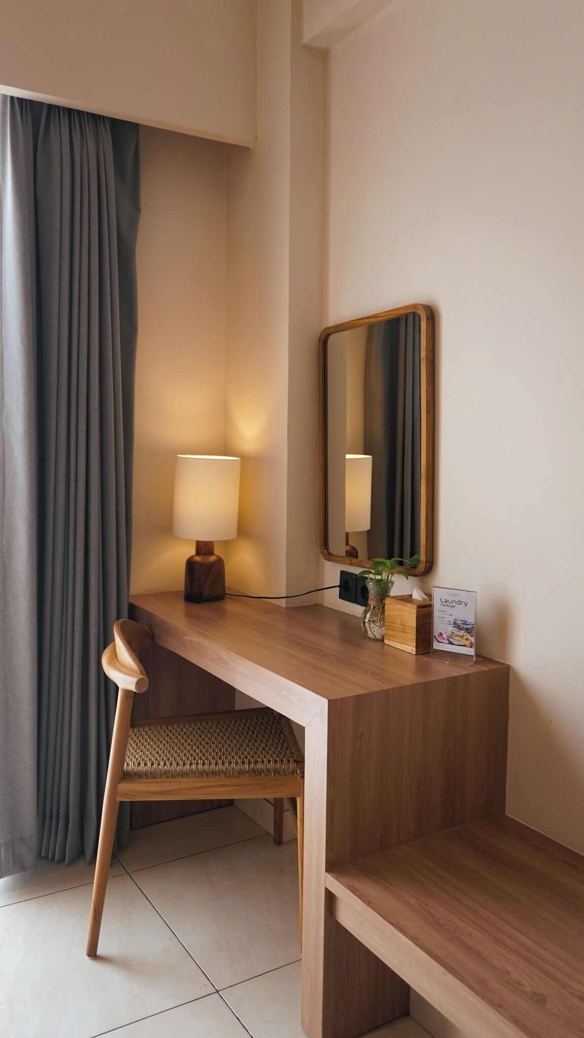A hotel room with a wooden desk, a chair, a lamp, a mirror, and a small plant.