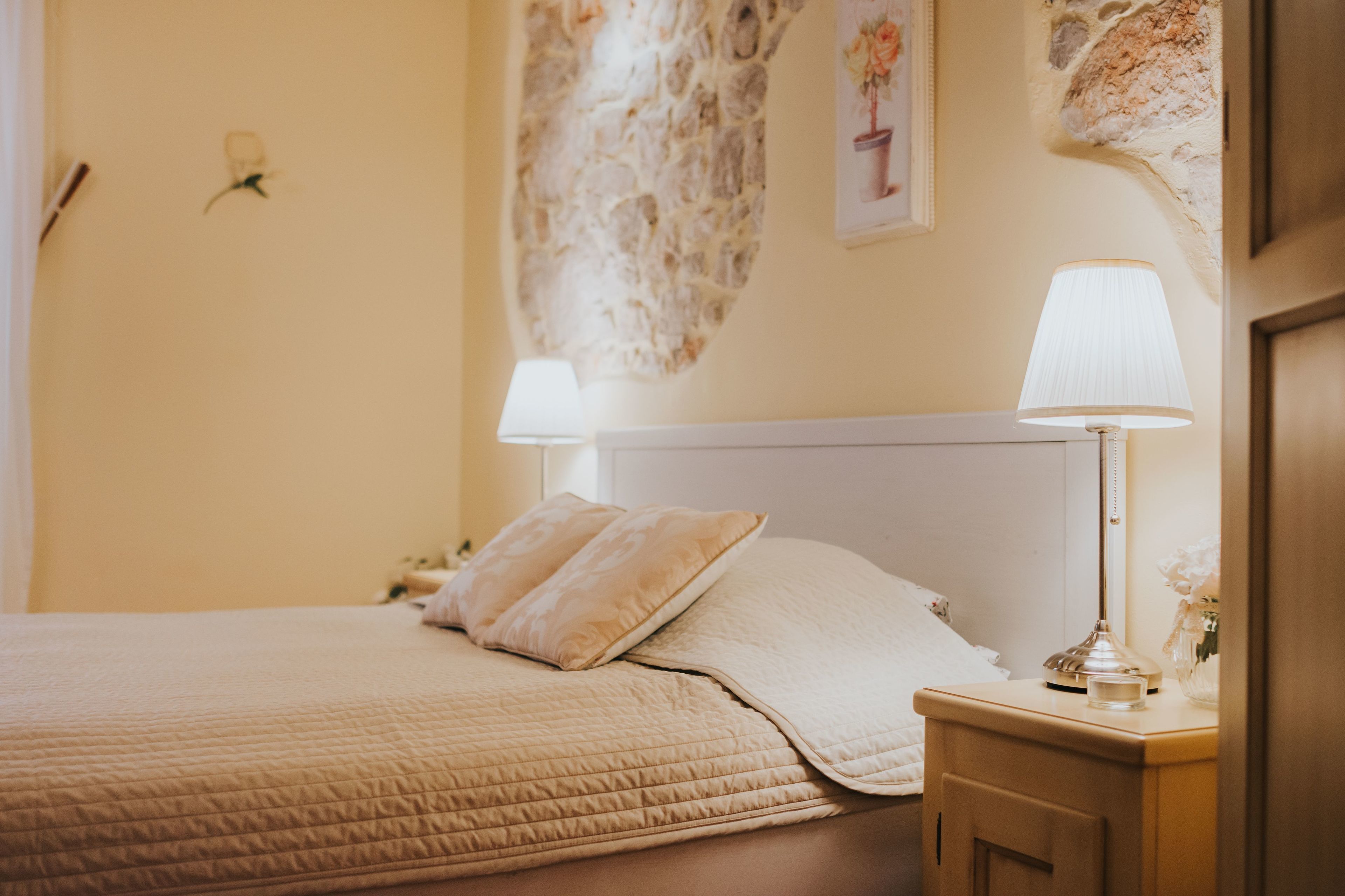 A neatly made bed with a beige quilt, a wooden nightstand with a lamp, and a stone wall with a floral painting.