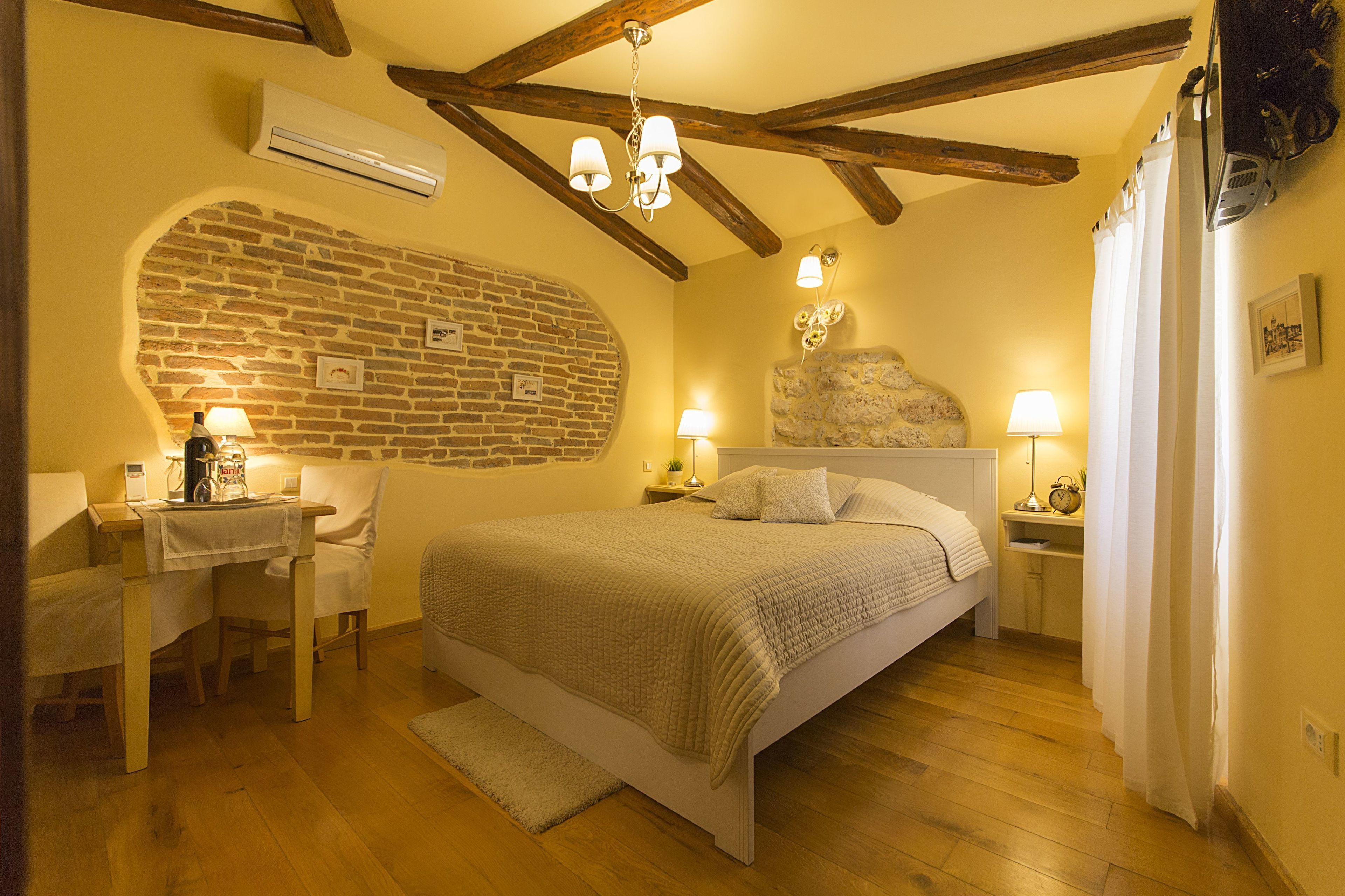 Un dormitorio con pared de ladrillo, vigas de madera, una cama, mesitas de noche con lámparas y un escritorio con silla.