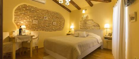 Un dormitorio con pared de ladrillo, vigas de madera, una cama, mesitas de noche con lámparas y un escritorio con silla.