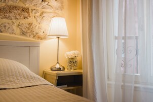 A neatly made bed with a white comforter, a bedside lamp, a vase with flowers, and a stone wall.