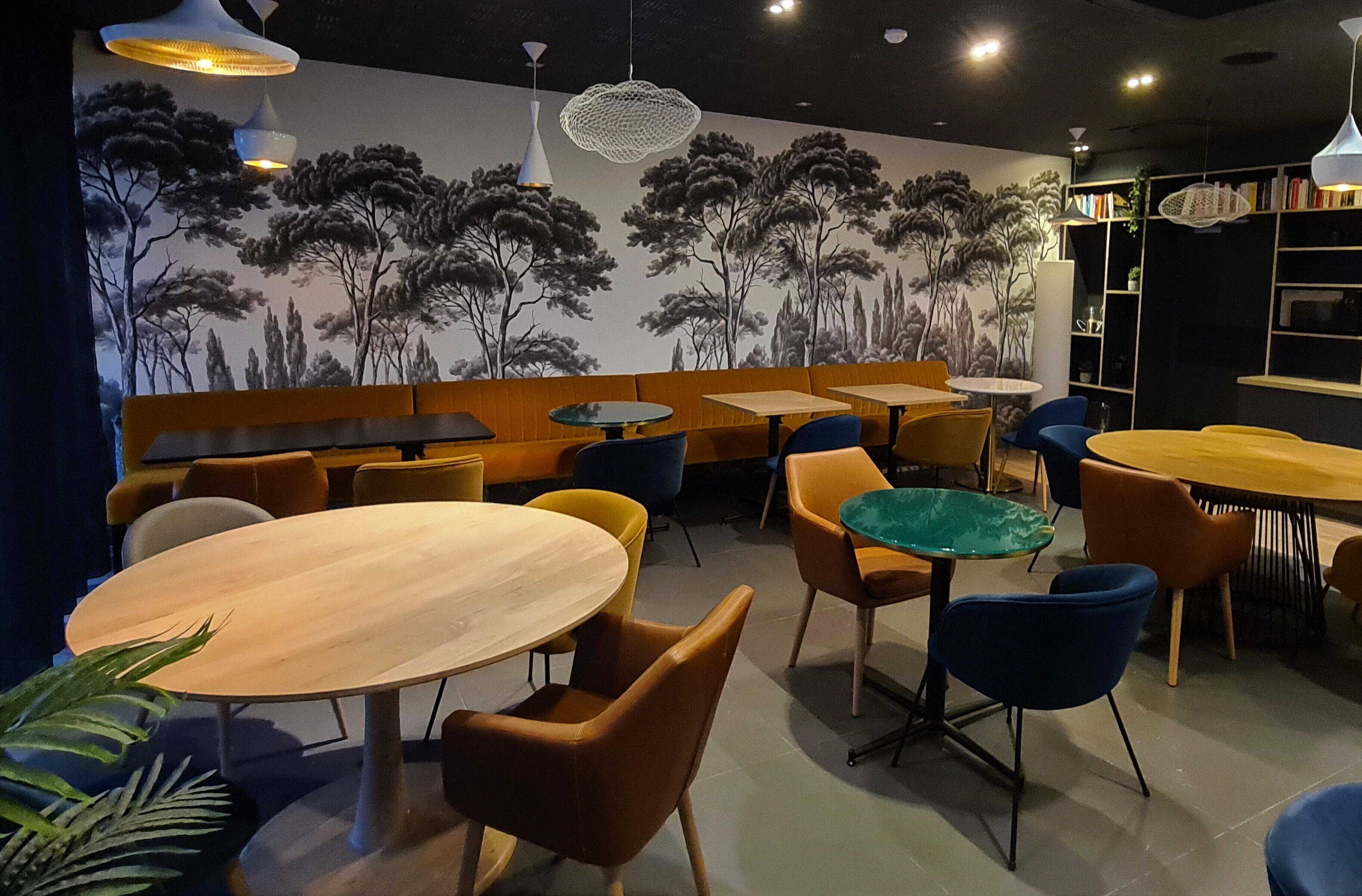 Un interior de restaurante con mesas redondas, sillas anaranjadas y azules, y un mural de árboles en la pared.
