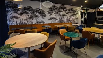 En restaurantindretning med runde borde, orange og blå stole og et muralmaleri af træer på væggen.