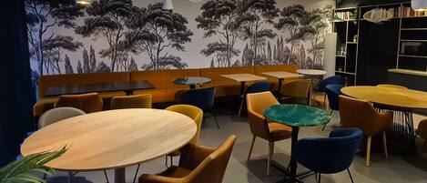A restaurant interior with round tables, orange and blue chairs, and a mural of trees on the wall.
