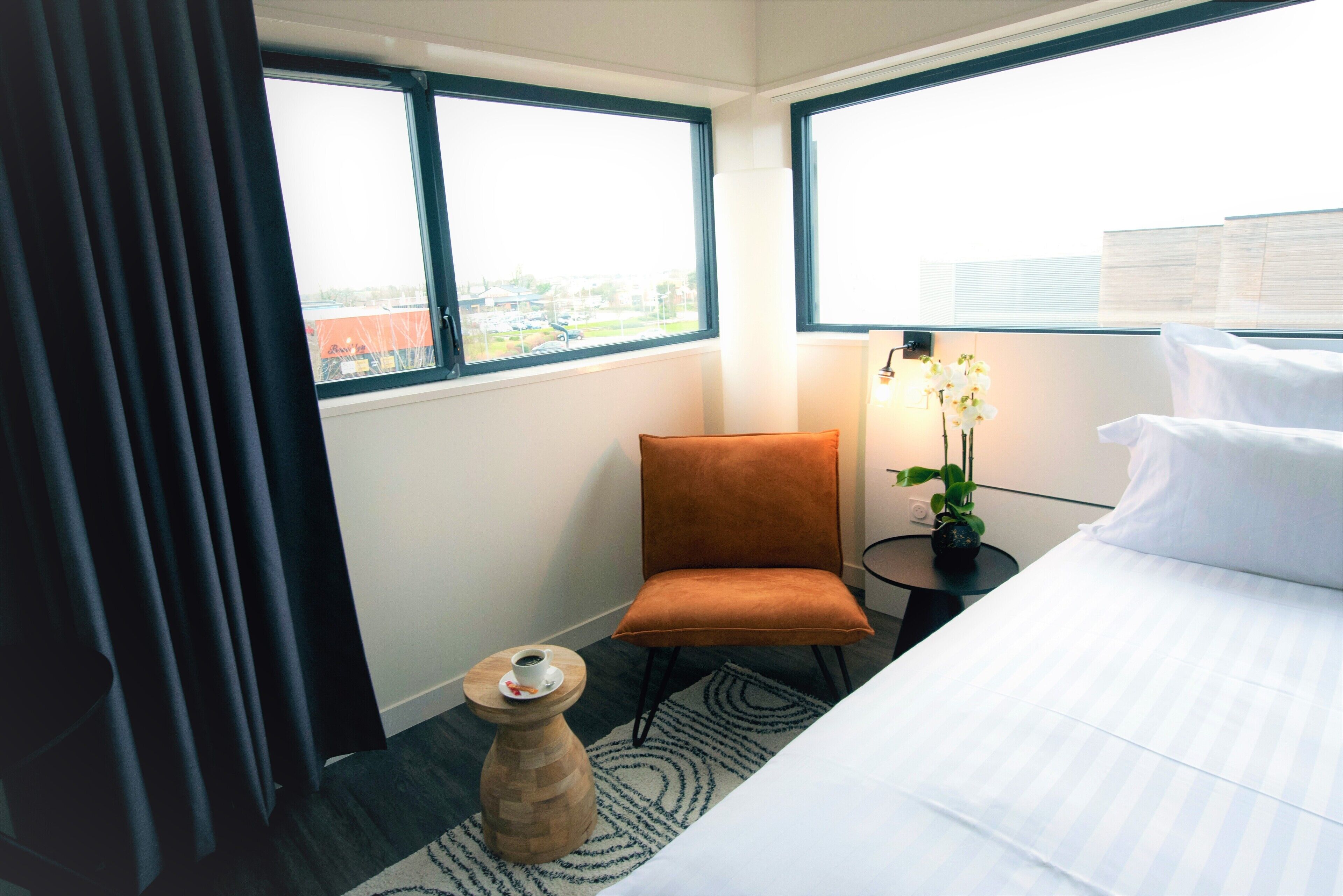 A hotel room with a bed, a small orange chair, a side table with a cup, a potted plant, and a view of the city through large windows.