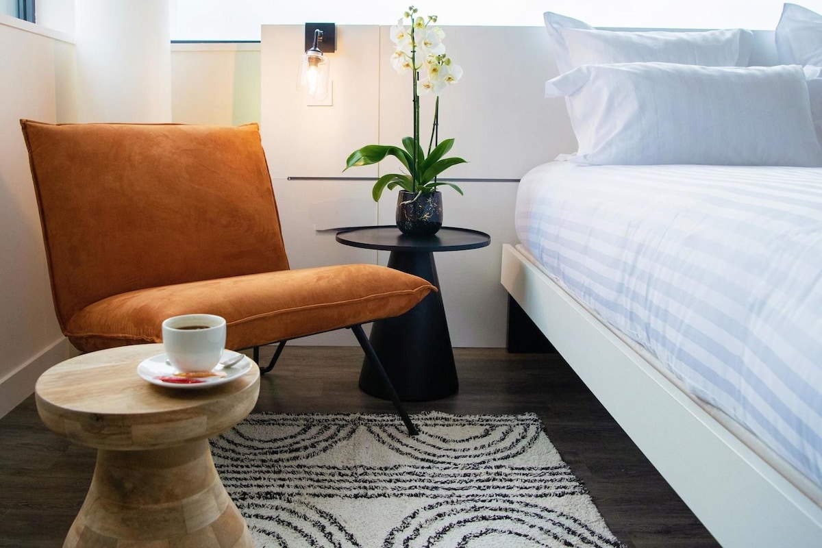 A hotel room with a bed, a chair, a small table with a cup, a potted plant, and a rug.