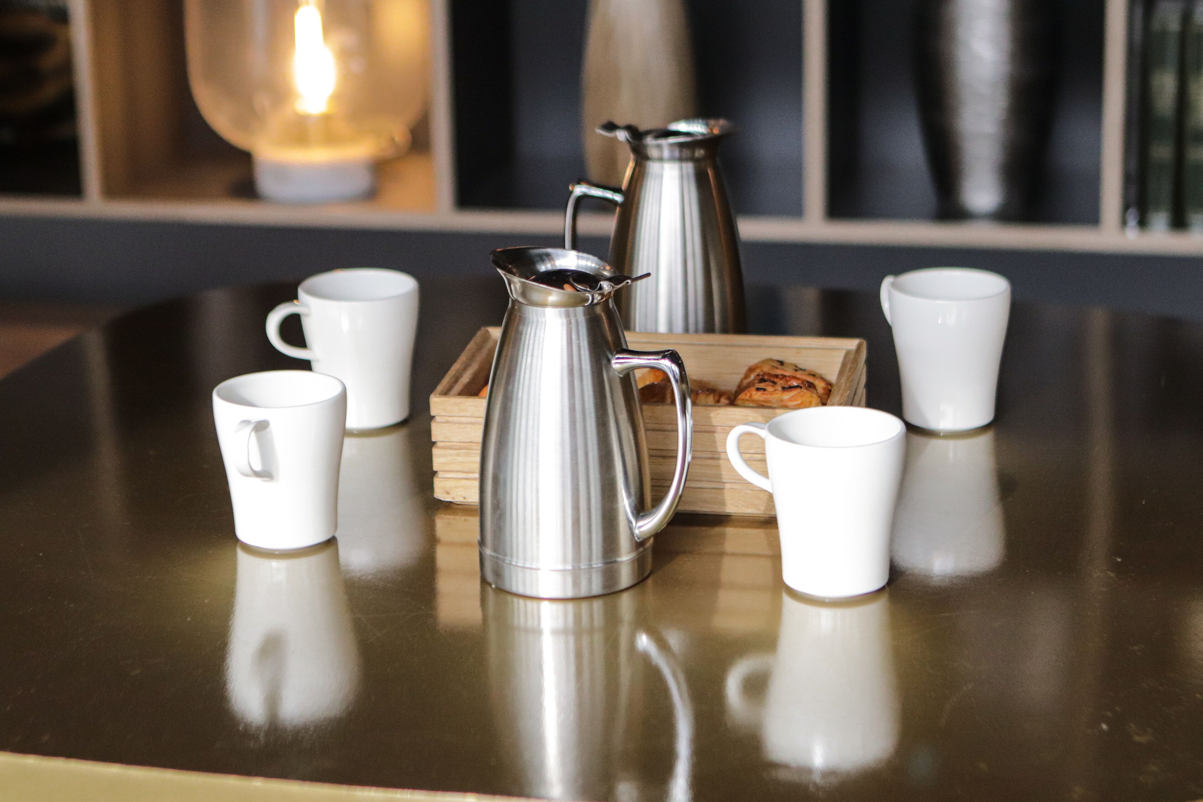A table with a metal coffee pot, a wooden tray with pastries, and several white cups.