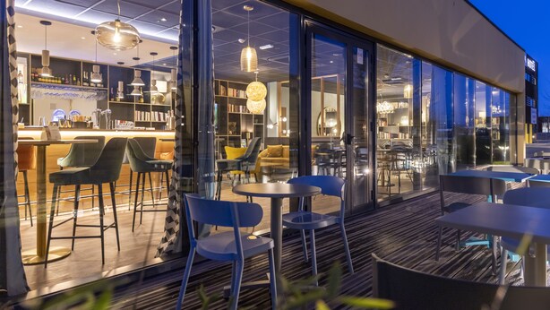 An indoor/outdoor seating area with tables and chairs, a bar area with shelves and bottles, and a view through large glass doors. - Padja Hotel & Spa, Vannes (Vannes)